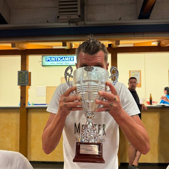 Ein Mann hält einen Pokal vor sein Gesicht und trägt ein weißes T-Shirt mit dem Aufdruck 'Meister'. Hinter ihm steht ein weiterer Mann an einer Theke, und ein blaues Schild mit 'Puntigamer' ist sichtbar.