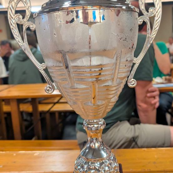 Ein glänzender silberner Pokal steht auf einem Holztisch. Er hat komplizierte Verzierungen auf seinem Körper. Dahinter sitzen Menschen auf Bänken, einige von ihnen tragen grüne Hemden.