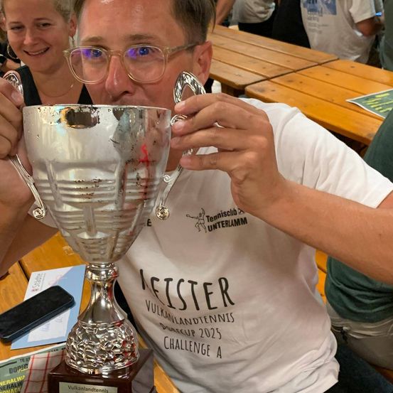 Ein Mann trinkt aus einer Trophäe, während er an einem Tisch sitzt, mit einer lächelnden Frau hinter ihm. Hinter ihnen sitzen Leute an einem Holztisch.