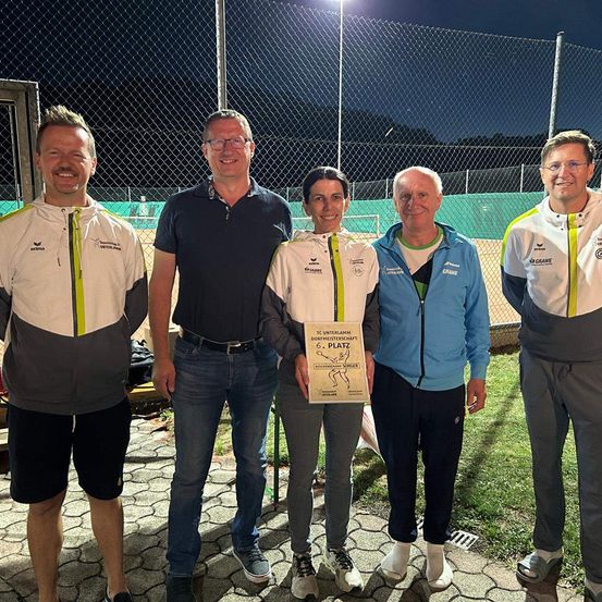 Fünf Erwachsene stehen draußen auf einem Rasenfeld. Eine Frau hält eine Plakette mit einem Bild einer Person darauf. Die Männer tragen Sportjacken. Hinter ihnen steht ein Metallzaun.