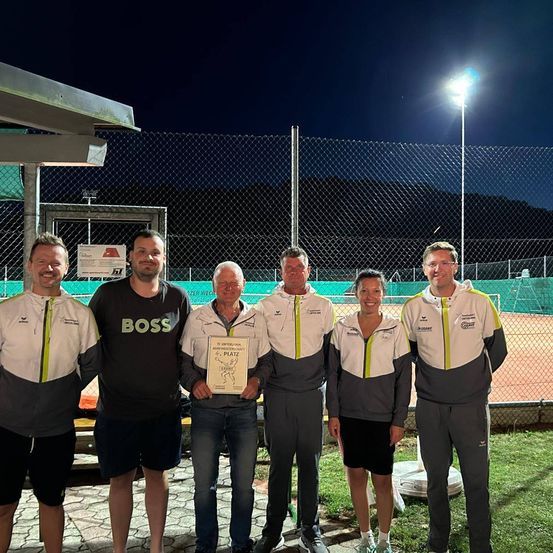 Fünf Personen stehen auf einem Tennisplatz, einer hält ein Zertifikat. Sie tragen passende Jacken, eine mit dem Wort Boss. Dahinter befindet sich ein Zaun und eine Lampe beleuchtet das Gelände.