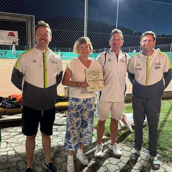 Drei Menschen stehen nebeneinander auf einem Grasfeld, wahrscheinlich auf einem Tennisplatz. Die Frau hält eine Urkunde mit gedrucktem Text. Der Mann links trägt einen weißen und grauen Mantel. Der Mann rechts trägt einen weißen Mantel. Der Mann in der Mitte trägt einen weißen Mantel und eine Hose.