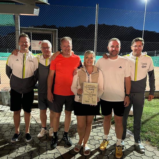 Fünf Personen stehen nachts auf einem Tennisplatz und lächeln. Eine Frau hält ein Zertifikat in der Mitte. Hinter ihnen steht ein Metallzaun, und in der Ferne sind Berge zu sehen.