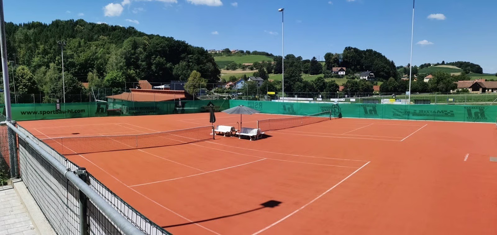 Ein Tennisplatz mit rotem Lehmboden, zwei Bänken, einem Regenschirm und einer Straßenlaterne unter einem blauen Himmel mit einigen Wolken und Grün im Hintergrund.
