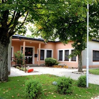 Ein Kindergartengebäude mit einem Schild, das 'Kindergarten Cvarnica' besagt. Das Gebäude hat eine weiße Außenfassade mit rosa Akzenten. Ein Fahnenmast steht im Vordergrund.