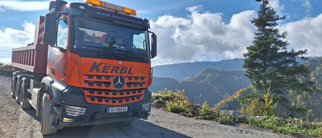 Ein Kerbl Schwerlastwagen fährt auf einer Bergstraße, mit Bergen im Hintergrund unter einem teilweise bewölkten Himmel.