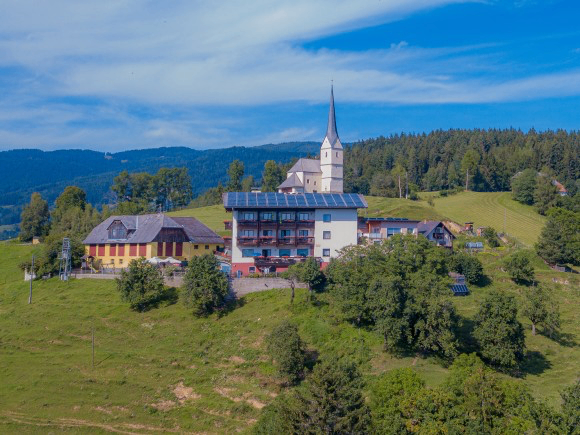 Luftaufnahme eines kleinen Dorfes mit einer Kirche und modernen Gebäuden, umgeben von üppigen grünen Feldern und Bäumen.