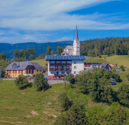 Luftaufnahme eines Dorfes mit einer Kirche und Häusern, umgeben von grünen Feldern und Bäumen. Die Kirche hat einen Turm und Solarpanele auf ihrem Dach.