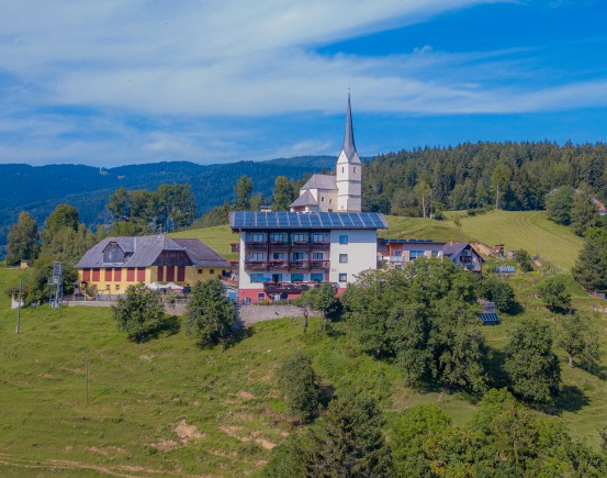 Luftaufnahme eines Dorfes mit einer Kirche und Häusern, umgeben von grünen Feldern und Bäumen. Die Kirche hat einen Turm und Solarpanele auf ihrem Dach.