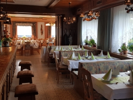 Ein leeres Restaurant mit Holztischen und Stühlen, weißen Tischdecken und grünen Servietten. Der Raum hat weiße Vorhänge, Holzböden und mehrere Lampen. Es gibt auch Topfpflanzen auf der Fensterbank.