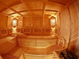 Ein holzverkleideter Saunaraum mit Holzbänken, einer Uhr an der Wand und zwei beleuchteten Fenstern. Der Raum ist von warmem, goldenem Licht durchflutet.