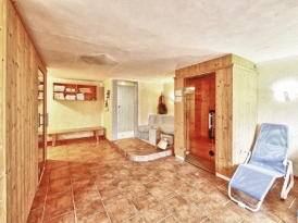 Ein gemütlicher Innensaunaraum mit Holzwänden, einer Holzbank und einem blauen Liegestuhl. Der Raum hat einen gefliesten Boden und einen Holzschrank mit Regalen.