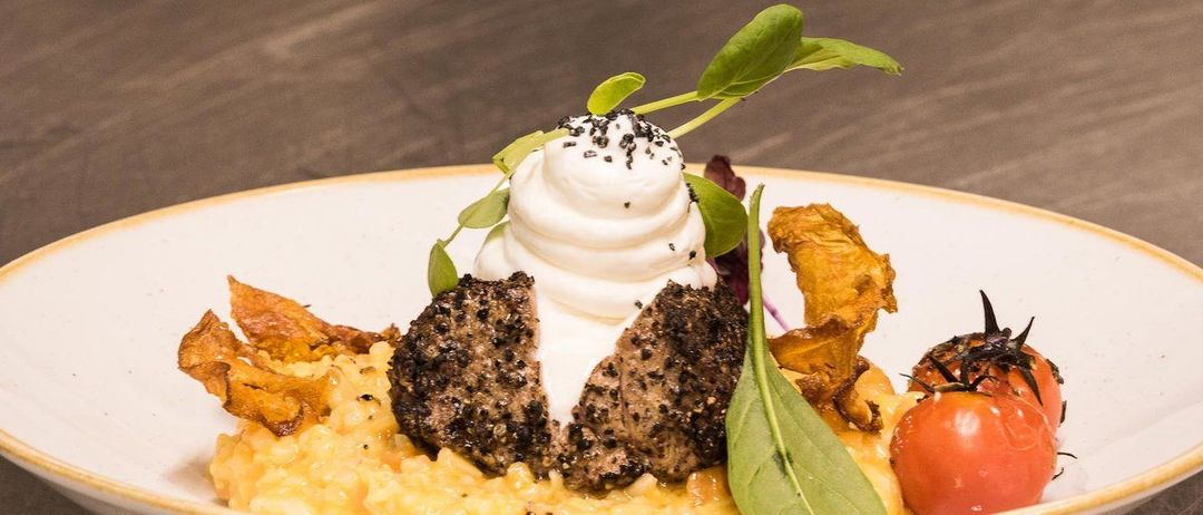 Ein Teller mit reichhaltigem, dunklem Fleisch, gekrönt von Schlagsahne und mit Grünzeug garniert, begleitet von cremigem Risotto, knusprigem Speck und Kirschtomaten.