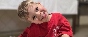 Ein junger Junge, der ein rotes T-Shirt trägt, posiert fröhlich mit einem geflochtenen Teig auf einem Backblech in einem Raum.