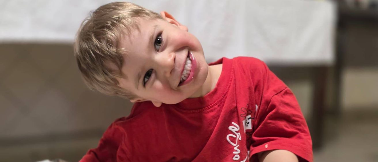 Ein junger Junge, der ein rotes T-Shirt trägt, posiert fröhlich mit einem geflochtenen Teig auf einem Backblech in einem Raum.