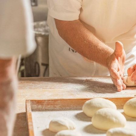 Ein Koch in einer Küche rollt Teig auf einem Holztisch. Mehrere Teigkugeln liegen auf einem Tablett vor ihm.