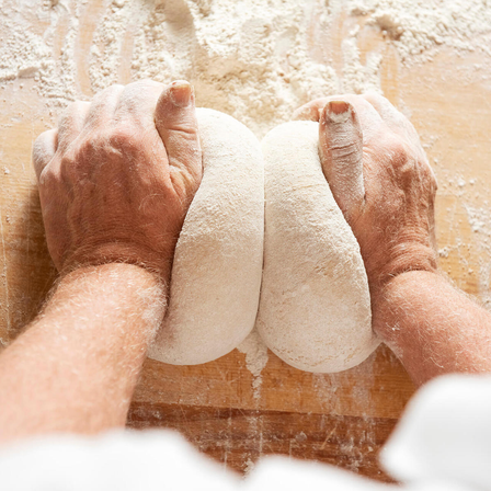 Hände kneten zwei Teigkugeln auf einer mehligen Holzfläche. Eine Hand liegt auf jeder Kugel und drückt nach unten.