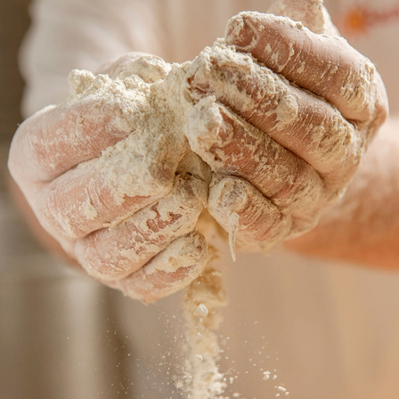 Mehlbestäubte Hände kneten Teig mit einem Hauch von Mehl, das abfällt. Die Person scheint Brot zuzubereiten.