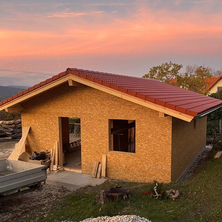 Ein kleines, unfertiges Haus mit rotem Dach steht auf einem Hang. Ein Lkw ist in der Nähe geparkt, und es gibt Haufen von Holz und anderen Materialien.