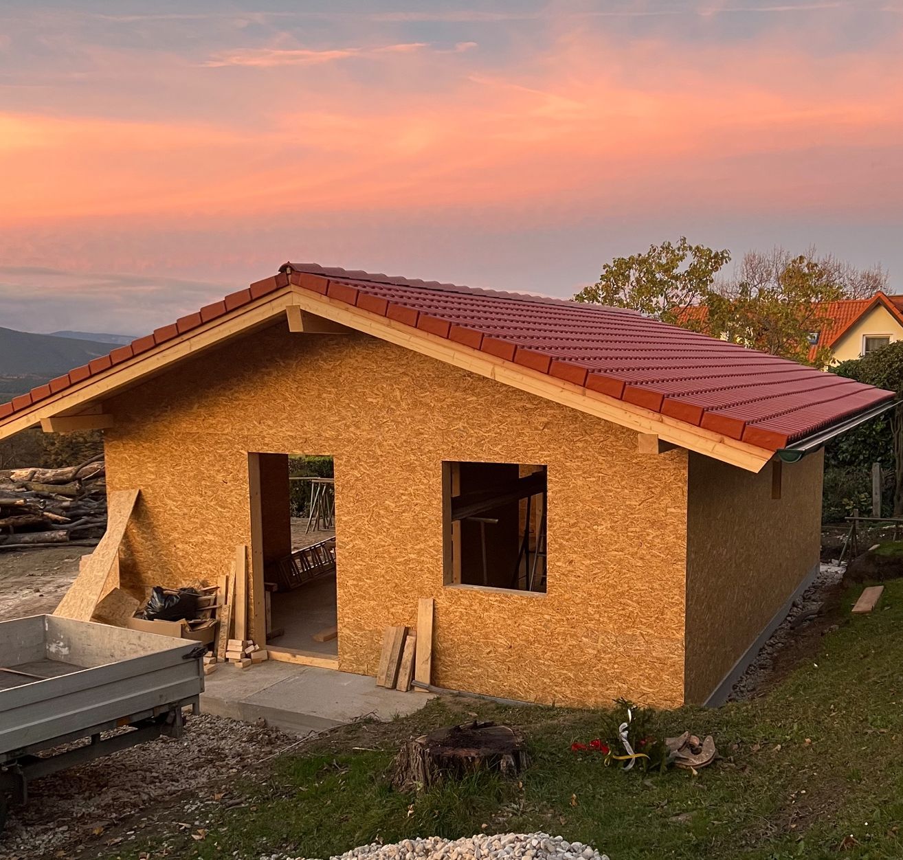 Ein kleines, unfertiges Haus mit rotem Dach steht auf einem Hang. Ein Lkw ist in der Nähe geparkt, und es gibt Haufen von Holz und anderen Materialien.