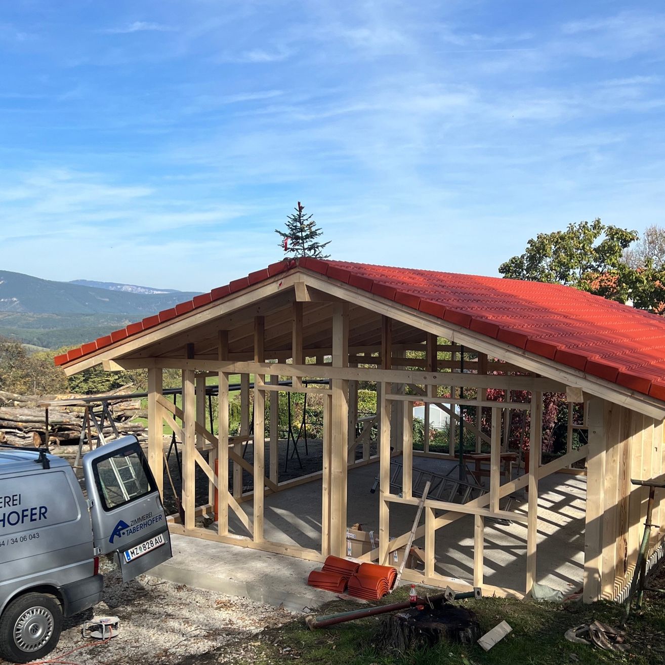 Ein Schuppen im Bau mit einem roten Dach. Er befindet sich in einem ländlichen Gebiet mit Bergen im Hintergrund. Ein Van ist neben dem Schuppen geparkt.