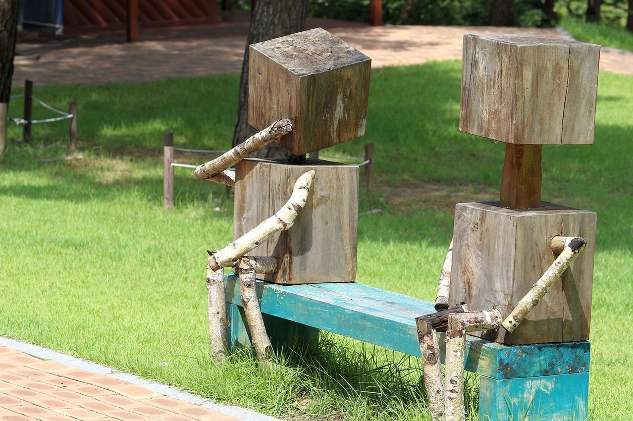 Two wooden sculptures resembling humans sit on a bench in a park. The figures have arms and legs made from twigs and branches.