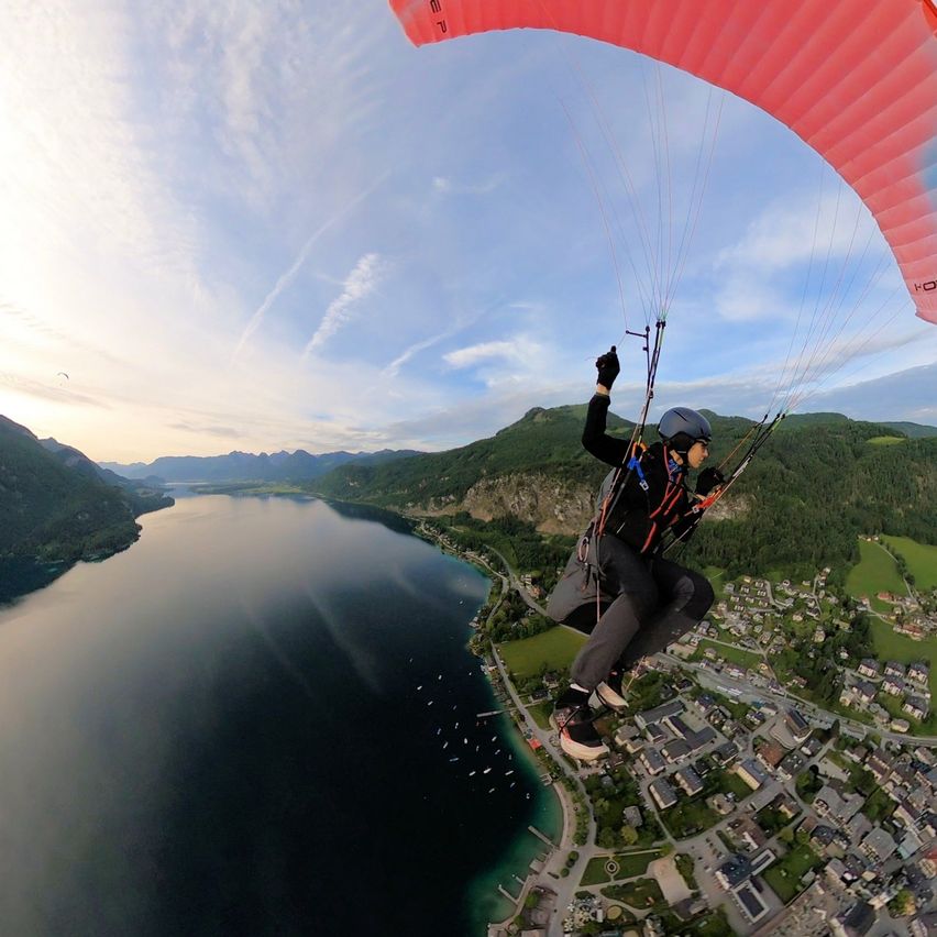 Eine Person paragliding über einer malerischen Landschaft mit Bergen, einem See und einer Stadt darunter. Der Himmel ist klar mit einigen Wolken.