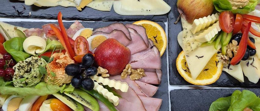 Eine Vielzahl von Lebensmitteln ist auf schwarzen Tellern angeordnet. Jeder Teller enthält verschiedene Zutaten wie Obst, Gemüse, Käse und Fleisch. Einige Teller enthalten geschnittenen Schinken, Orangenscheiben und andere Zutaten.