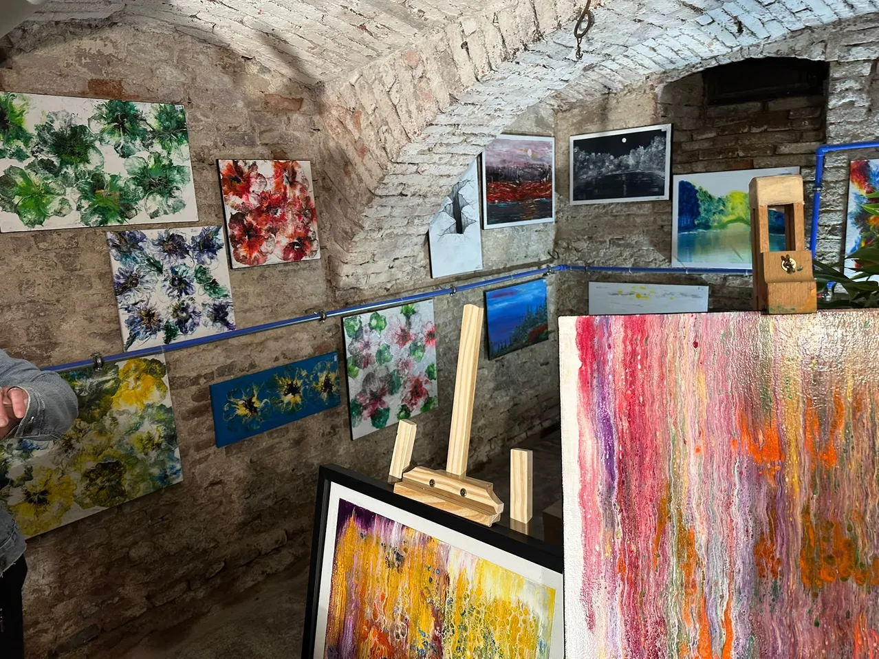 Eine Kunstgalerie mit Ziegelwänden zeigt verschiedene abstrakte Gemälde. Die Gemälde sind lebendig und farbenfroh, hängen an den Wänden und werden von Holzstaffeln gestützt.