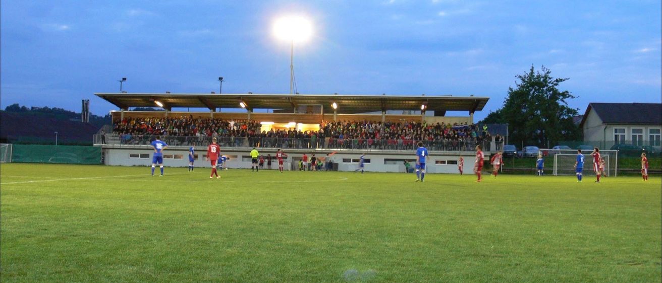 Ein Fußballspiel findet bei Nacht unter Flutlichtern statt, mit Spielern in roten und blauen Uniformen auf dem Feld und Zuschauern in den Tribünen.