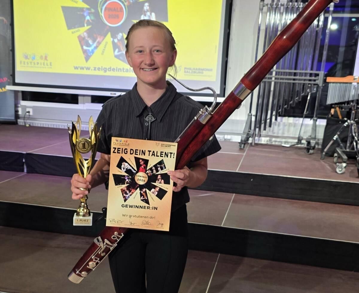 A young girl holding a golden trophy and a poster with a text 'Zeige dein Talent' on it, standing on a stage. She is smiling and holding a bassoon. Behind her is a large screen with some text and logos.