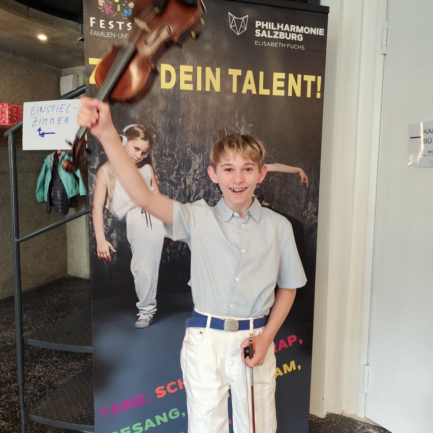 A boy stands in front of a poster holding a violin. The poster says 'Your Talent!' with a girl dancing in the background.