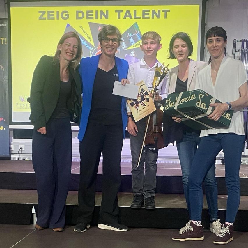 Five people stand on stage, smiling for the camera. A boy holds a violin and award, a woman holds a box. A banner behind them reads 'Zeige Dein Talent.'