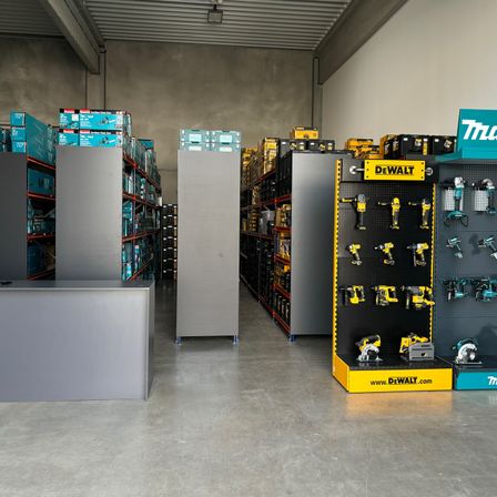 Ein Lagerraum-Interieur mit mehreren Werkzeugdisplays und Lagerregalen. DEWALT- und Makita-Werkzeuge sind auf schwarzen und gelben Ständern prominent ausgestellt.