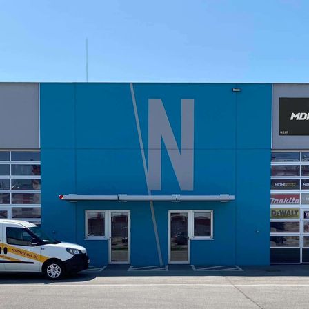 Ein blaues Gebäude mit einem großen 'N' auf der Vorderseite. Vor dem Gebäude steht ein weißer Lieferwagen. Das Gebäude hat Glasfenster und Türen.