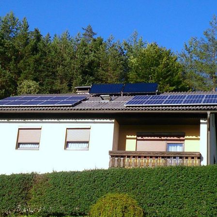 Ein Haus mit Solarpanelen auf dem Dach, umgeben von Büschen und Bäumen unter einem klaren blauen Himmel.