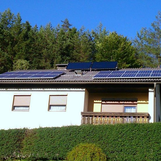 Ein Haus mit Solarpanelen auf dem Dach, umgeben von Büschen und Bäumen unter einem klaren blauen Himmel.