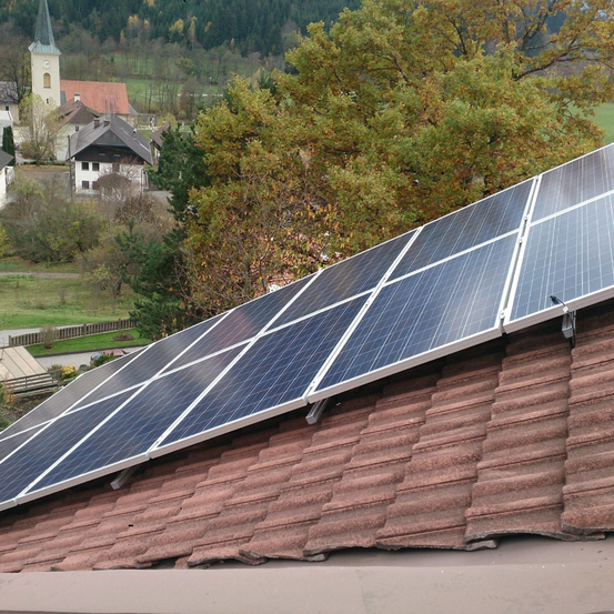 Ein Wohndach mit mehreren Solarmodulen. Dahinter befindet sich ein kleines Dorf mit einer Kirche und mehreren Häusern.