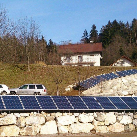 Ein Haus mit einem braunen Dach steht auf einem Hügel mit Bäumen. Zwei Autos sind in der Nähe von Solarpanelen geparkt. Die Solarpanele sind auf einer Steinmauer angebracht.