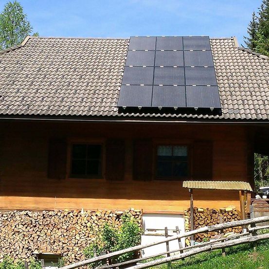 Ein Holzhaus mit Solarpanelen auf dem Dach, einem Schuppen und gestapeltem Brennholz bei sonnigem Wetter.