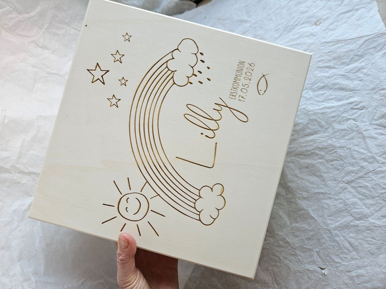 A wooden box with a rainbow, stars, and sun on its cover. The name 'Lilly' and the date '17.05.2026' are engraved on the cover. A hand is holding the box.