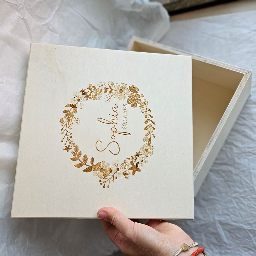 A hand holds a white box with a floral wreath around the name 'Sophia' and the date '30.07.2005'. The box is placed on a white surface.