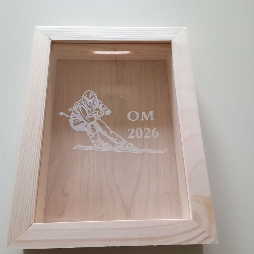 Eine Holzbox mit Glasfront zeigt einen Skifahrer in Aktion, beschriftet mit OM 2026. Der Skifahrer ist inmitten der Abfahrt festgehalten und zeigt die Schönheit des Skifahrens.