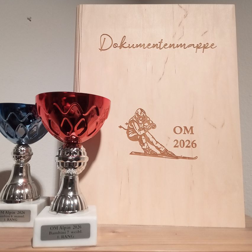 Zwei Trophäen mit einem roten und blauen Oberteil werden mit einer Holzbox mit Gravuren ausgestellt. Die Trophäen haben ein Etikett mit dem Text OM Alpin 2026 Bambini 7 weibl. 1 RANG. Die Box hat Gravuren eines Skifahrers und den Text Dokumentenmappe OM 2026.