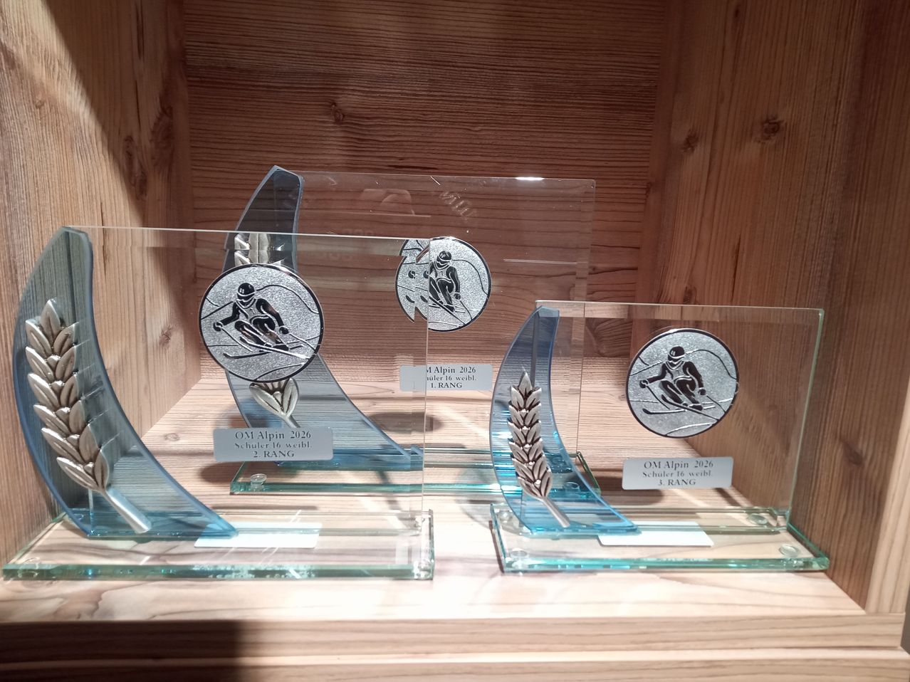 Drei Glastrophäen auf einem Holzregal. Jede Trophäe zeigt ein Skifahrerdesign und Text. Die erste Trophäe sagt 'OM Alpin 2026 Schüler 16 weib 2 RANG'. Die zweite Trophäe sagt '4 Alpin 2026 Schüler 16 weib 1 RANG'. Die dritte Trophäe sagt 'OM Alpin 2026 Schüler 16 weib 3 RANG'.