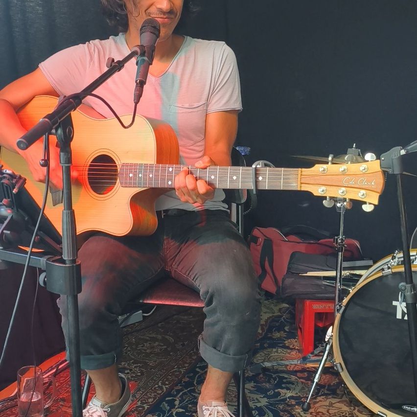 Ein Mann sitzt auf einem Stuhl und spielt Gitarre. Ein Mikrofon steht vor ihm. Ein Schlagzeug und eine Tasche befinden sich hinter ihm.