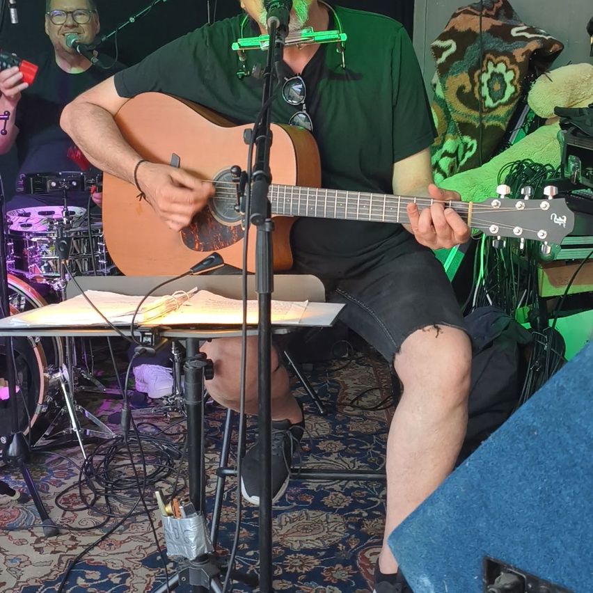 Ein Mann spielt Gitarre mit einem Mundharmonika nahe einem Schlagzeug. Er sitzt mit einem Notenständer vor sich, während ein anderer Mann mit Brille in der Nähe singt.