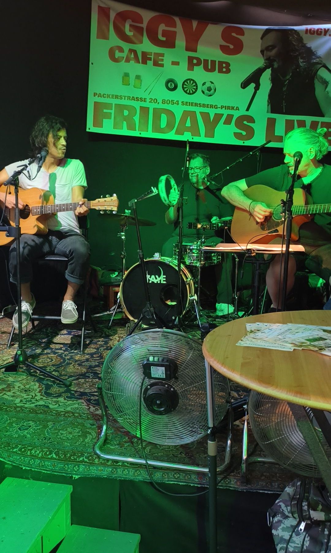 Drei Musiker treten auf der Bühne auf. Ein Banner mit der Aufschrift Friday's Live hängt hinter ihnen. Die Musiker spielen Gitarre, Schlagzeug und Becken. Ein Ventilator und ein Tisch mit Papieren stehen vor ihnen.