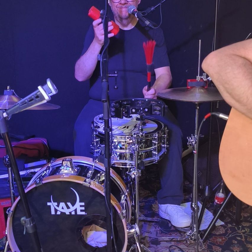 Ein Mann mit Brille und blauem T-Shirt sitzt und spielt ein Schlagzeug mit einem Mikrofon vor sich.