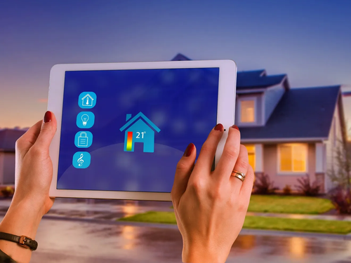 Hände halten ein Tablet mit Smart-Home-Steuerungen, einem Haussymbol und einer Temperaturanzeige von 21°. Ein Haus ist im Hintergrund.